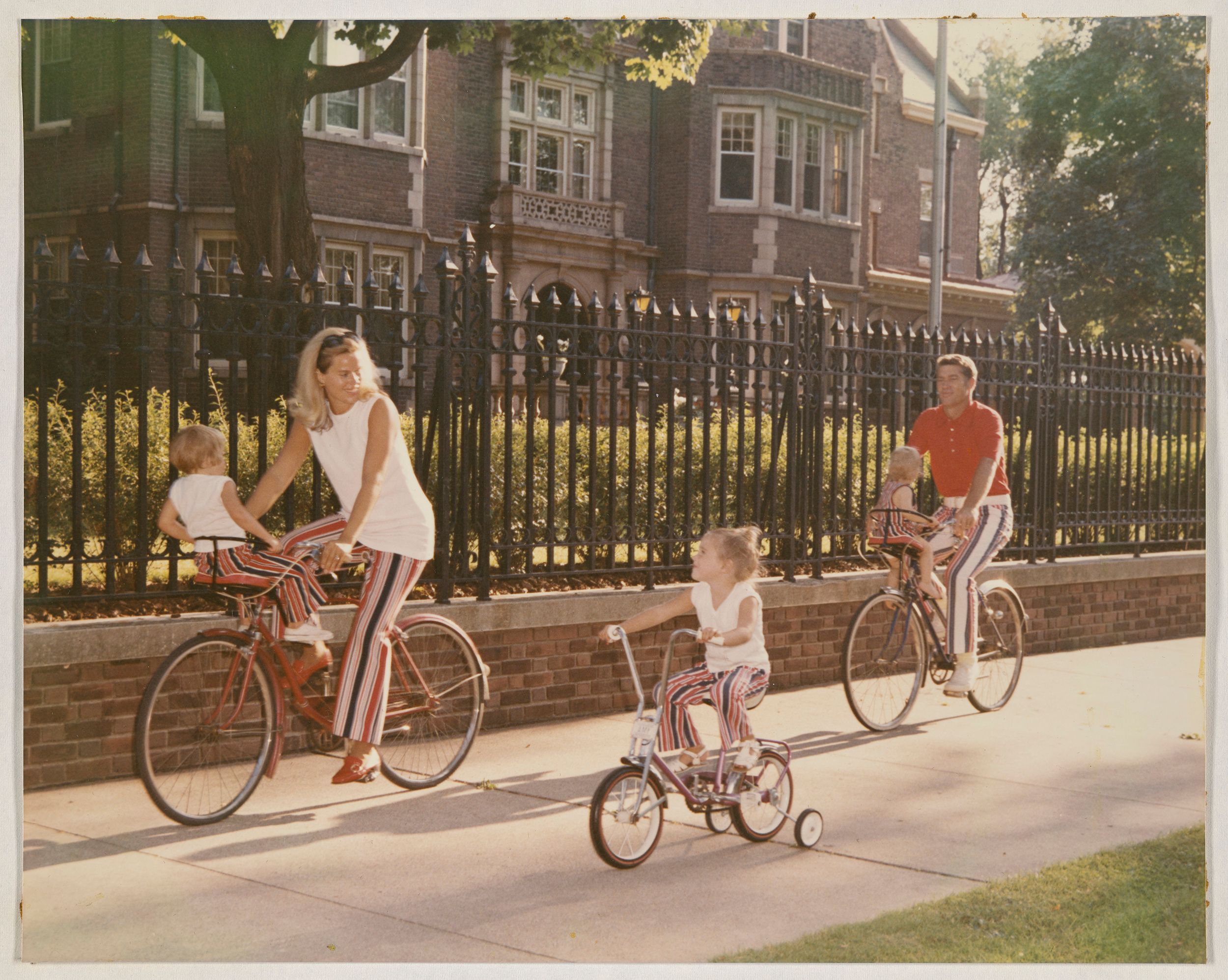 Family riding bikes on a sidewalk in front of a large brick building with iron fencing, all dressed in coordinated striped pants and white or red tops.